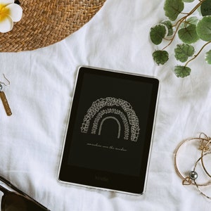 May include: A black and white illustration of a rainbow on a Kindle e-reader. The text "somewhere over the rainbow" is displayed below the rainbow. The Kindle is on a white surface with a straw hat, earrings, and a plant in the background.