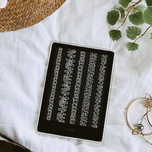 May include: A black and white Kindle e-reader displaying a page with five different floral border designs. The e-reader is on a white surface with a straw hat, earrings, and bracelets.