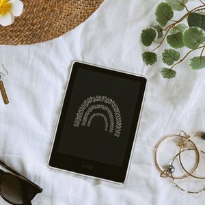 May include: A white background with a black Kindle e-reader displaying a rainbow design. The e-reader is surrounded by a straw hat, earrings, sunglasses, and a gold bracelet with a pearl.
