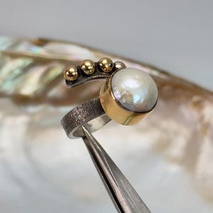 May include: A silver ring with a textured band and a large white pearl set in a gold bezel. The ring is accented with five small gold beads.
