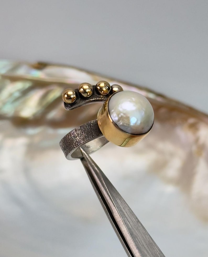 May include: A silver ring with a textured band and a large white pearl set in a gold bezel. The ring is accented with five small gold beads.