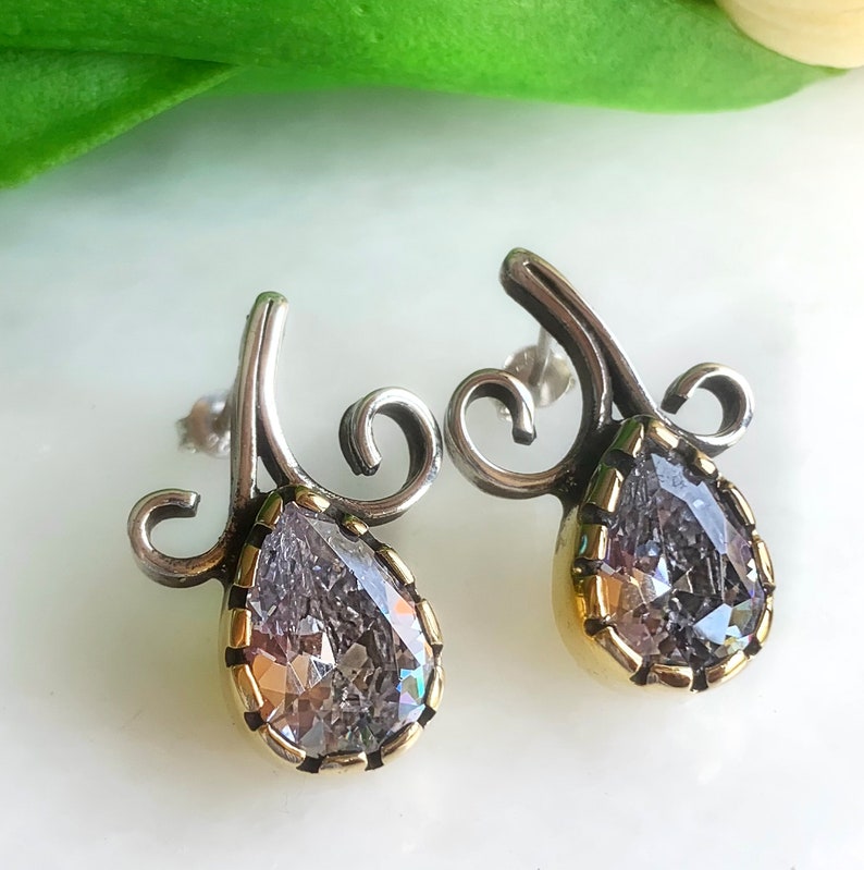 May include: A pair of silver earrings with a teardrop-shaped clear crystal set in a gold-toned bezel. The earrings feature a delicate, swirling design.
