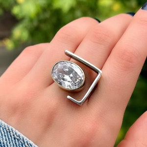 Puede incluir: Un anillo de plata y oro con una gran piedra ovalada transparente engastada en el centro. El anillo tiene un dise&ntilde;o geom&eacute;trico &uacute;nico con un marco cuadrado.