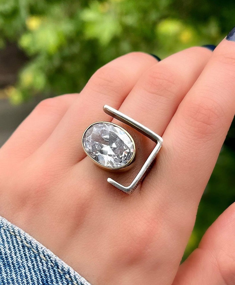 Puede incluir: Un anillo de plata y oro con una gran piedra ovalada transparente engastada en el centro. El anillo tiene un dise&ntilde;o geom&eacute;trico &uacute;nico con un marco cuadrado.