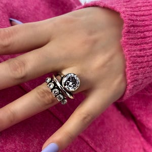 May include: A silver ring featuring a large, round, clear gemstone and a smaller ring with several smaller clear gemstones. The ring is worn on a hand with a pink sweater and light purple nail polish.