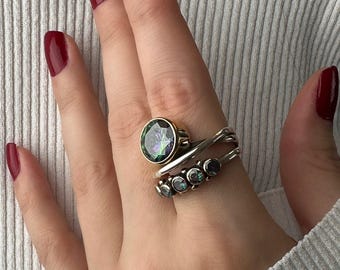 May include: A hand wearing three rings. The largest ring has a gold band and a large, round, iridescent stone. The second ring is silver with a crisscross design. The third ring is silver with five small iridescent stones.