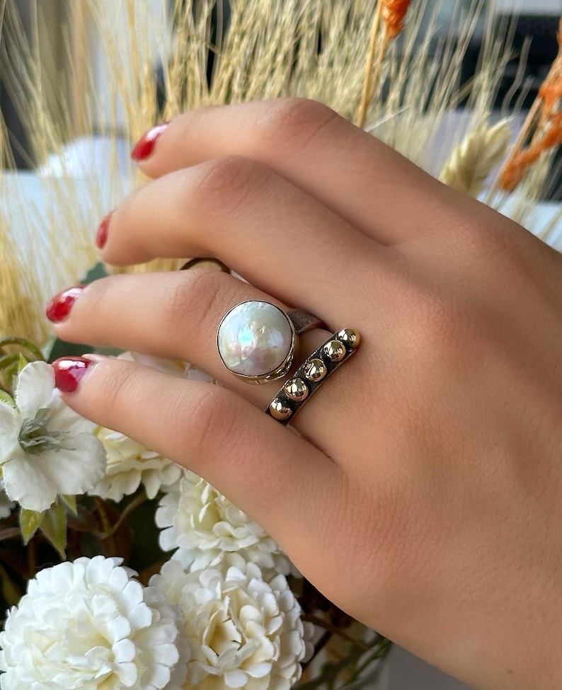 May include: A silver ring with a large white pearl and a gold band with small gold balls. The ring is on a hand with red nail polish.