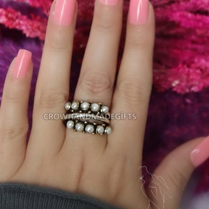 May include: A gold ring with a row of white pearls. The ring is worn on a hand with pink nail polish.