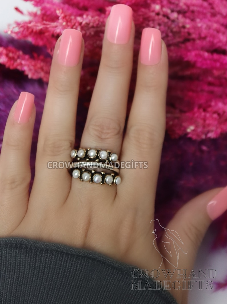 May include: A gold ring with a row of white pearls. The ring is worn on a hand with pink nail polish.