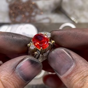 Könnte beinhalten: Ein gold-silberner Ring mit einem großen, oval geschliffenen, feuerroten Edelstein als Mittelpunkt. Der Ring ist mit kleineren, klaren Edelsteinen und aufwendigen Golddetails verziert. Der Ring wird zwischen zwei Händen gehalten.