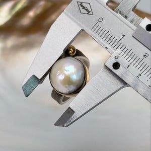 May include: A silver ring with a large, white pearl and a gold accent. The ring is being measured with a caliper, showing the pearl is approximately 1.5 cm in diameter.