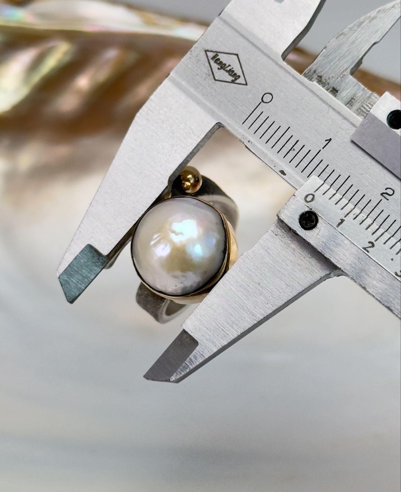 May include: A silver ring with a large, white pearl and a gold accent. The ring is being measured with a caliper, showing the pearl is approximately 1.5 cm in diameter.