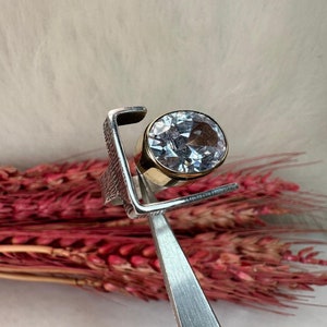 Puede incluir: Un anillo de plata con una gran piedra redonda transparente engastada en una banda de oro. El anillo est&aacute; dise&ntilde;ado con un marco cuadrado alrededor de la piedra preciosa.