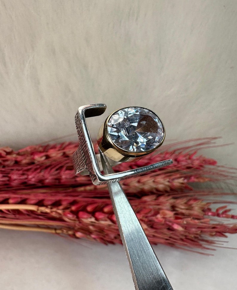 Puede incluir: Un anillo de plata con una gran piedra redonda transparente engastada en una banda de oro. El anillo est&aacute; dise&ntilde;ado con un marco cuadrado alrededor de la piedra preciosa.