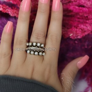 May include: A close-up of a hand wearing a ring with multiple bands of small, white pearls. The hand is painted with a light pink nail polish. The background is a pink, fuzzy material.