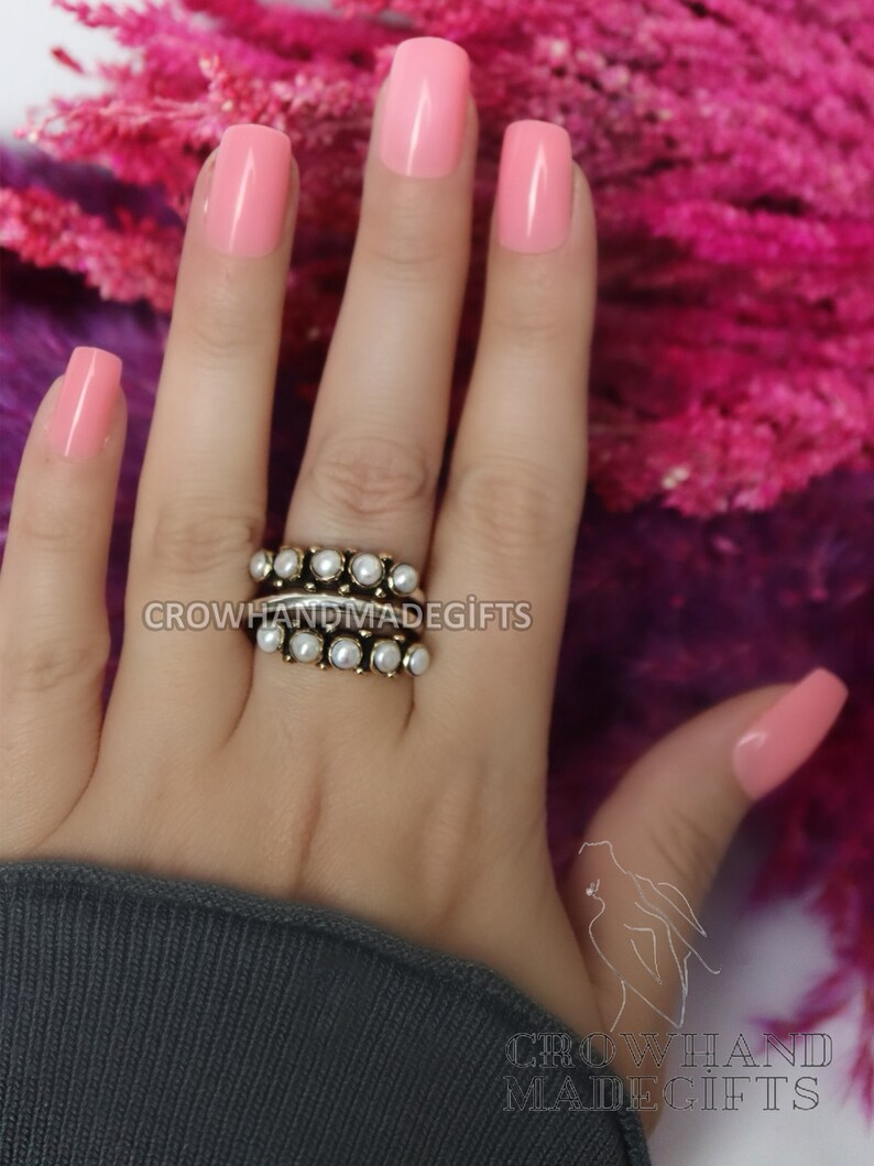 May include: A close-up of a hand wearing a ring with multiple bands of small, white pearls. The hand is painted with a light pink nail polish. The background is a pink, fuzzy material.