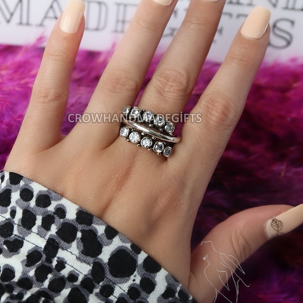 Puede incluir: Un anillo de plata con una fila de piedras de circonita cúbica transparentes. El anillo está en una mano con esmalte de uñas rosa claro. El texto "CROWHANDMADEGIFTS" es visible en la imagen.