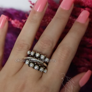 May include: A silver ring with a row of white pearls on each side of a silver band. The ring is worn on a finger with pink nail polish.