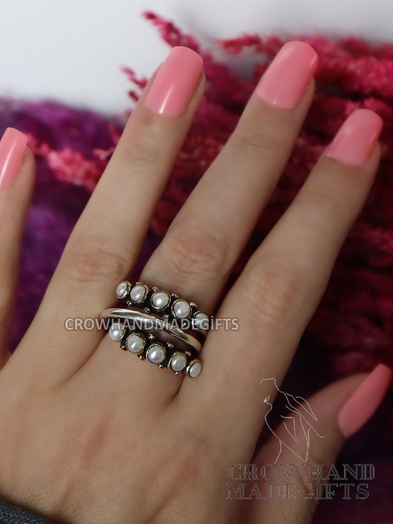 May include: A silver ring with a row of white pearls on each side of a silver band. The ring is worn on a finger with pink nail polish.