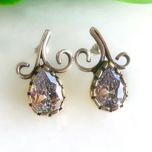May include: A pair of silver earrings with a pear-shaped clear crystal set in a gold-toned bezel. The earrings have a delicate, swirling design.