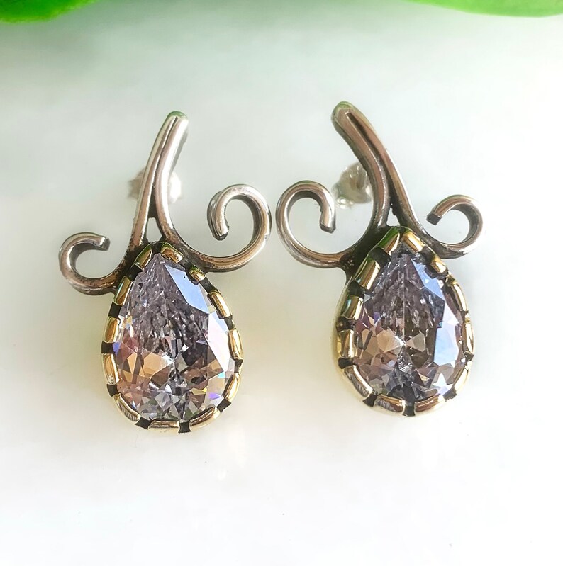 May include: A pair of silver earrings with a pear-shaped clear crystal set in a gold-toned bezel. The earrings have a delicate, swirling design.