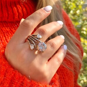 May include: A gold ring with a large, clear, round gemstone and smaller pear-shaped gemstones. The ring is on a finger wearing a red sweater.
