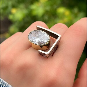 Puede incluir: Un anillo de plata con un engaste de oro y una gran piedra de circonita c&uacute;bica transparente. El anillo tiene un dise&ntilde;o geom&eacute;trico con una banda texturizada.