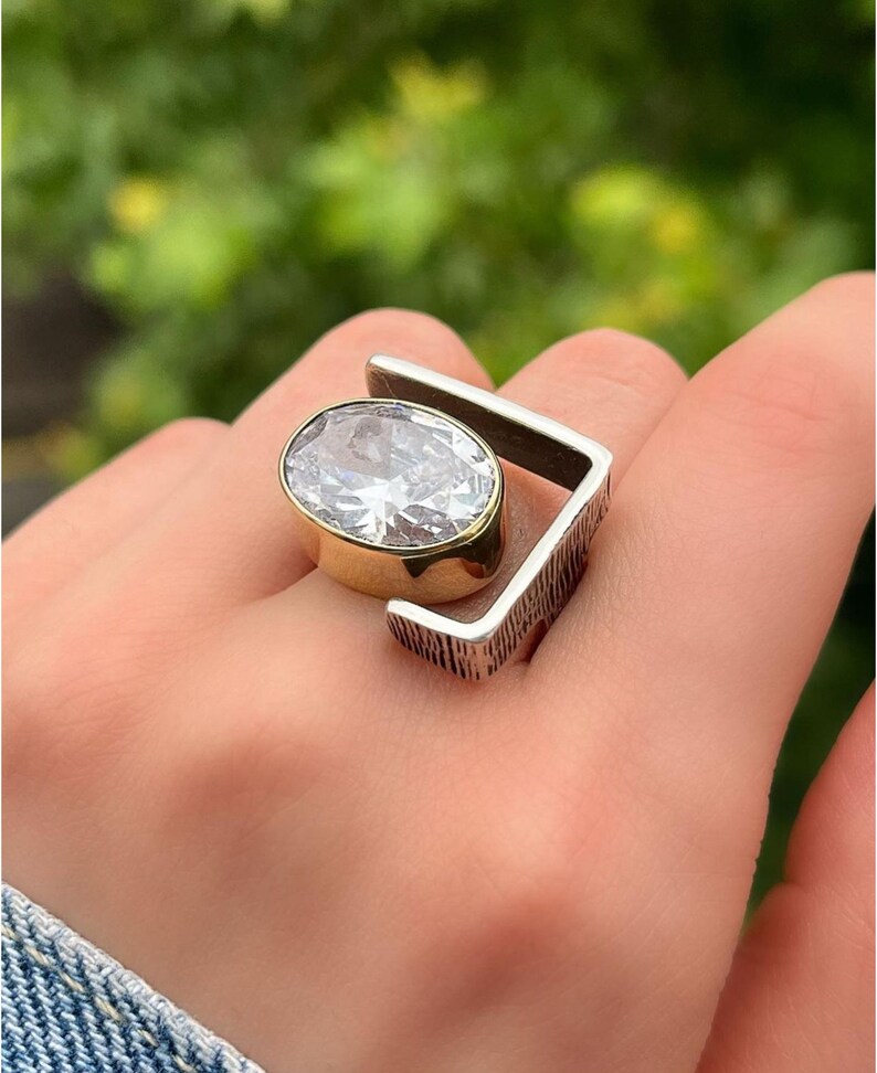 Puede incluir: Un anillo de plata con un engaste de oro y una gran piedra de circonita c&uacute;bica transparente. El anillo tiene un dise&ntilde;o geom&eacute;trico con una banda texturizada.