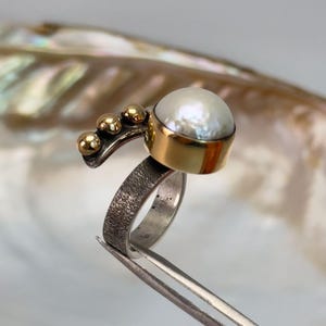 May include: A silver ring with a textured band and a gold setting featuring a white pearl. The ring has a gold accent with three small gold balls.