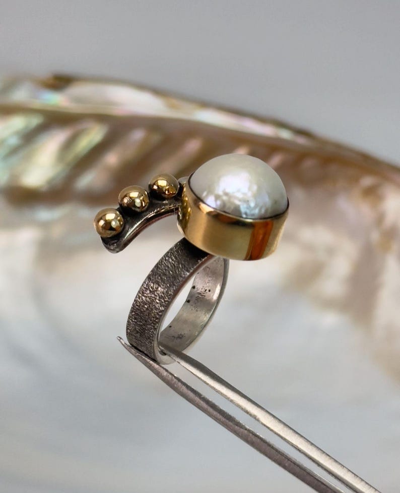 May include: A silver ring with a textured band and a gold setting featuring a white pearl. The ring has a gold accent with three small gold balls.