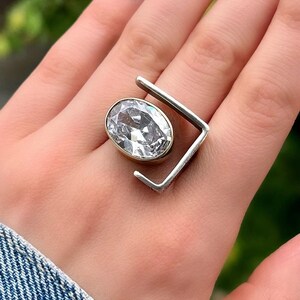 Puede incluir: Un anillo de plata con una gran piedra ovalada transparente engastada en un engaste de oro. El anillo tiene un dise&ntilde;o geom&eacute;trico &uacute;nico con un marco cuadrado.