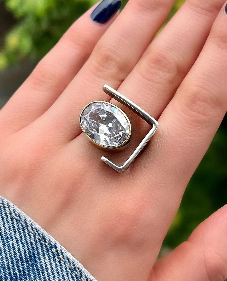 Puede incluir: Un anillo de plata con una gran piedra ovalada transparente engastada en un engaste de oro. El anillo tiene un dise&ntilde;o geom&eacute;trico &uacute;nico con un marco cuadrado.