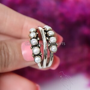May include: A silver ring with two bands, each adorned with a row of white pearls. The ring is held by a hand against a pink and purple background.