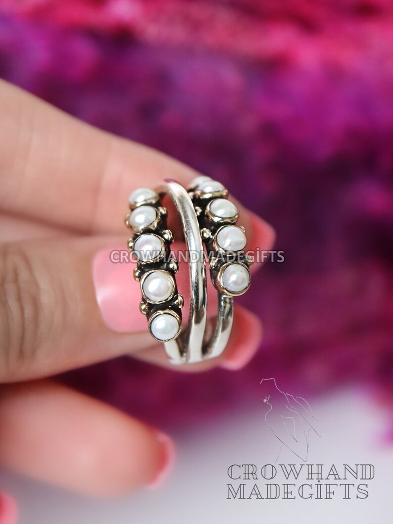 May include: A silver ring with two bands, each adorned with a row of white pearls. The ring is held by a hand against a pink and purple background.
