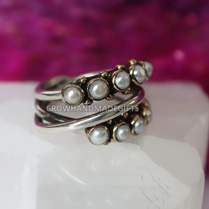 May include: A silver and gold ring with a row of white pearls. The ring is on a white surface.  CROWHANDMADEGIFTS is printed on the ring.