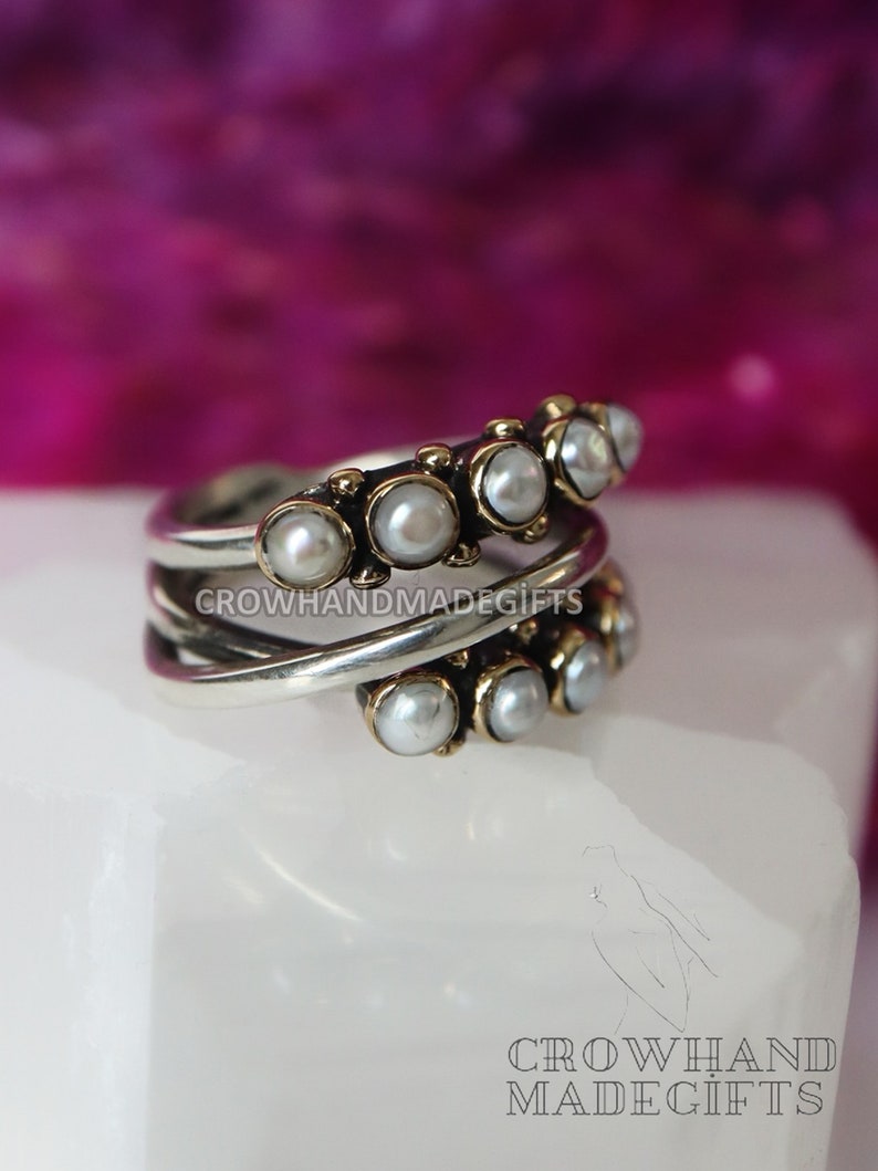 May include: A silver and gold ring with a row of white pearls. The ring is on a white surface.  CROWHANDMADEGIFTS is printed on the ring.