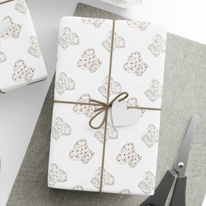 May include: Three white gift boxes wrapped in brown twine and decorated with a white and brown pattern of gingerbread cookies. The boxes are on a white surface with a gray fabric background.