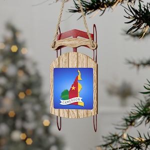 Pode incluir: Um ornamento de Natal de madeira em forma de trenó com a parte superior vermelha. O trenó tem um fundo azul com um mapa de Camarões e a palavra "Camarões" em uma fita branca.