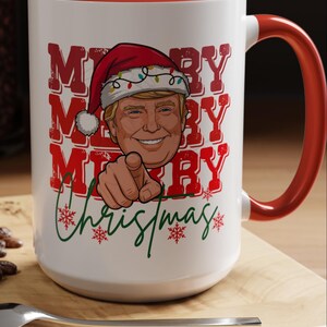 May include: White ceramic mug with a red interior and handle. The mug features a graphic of a person wearing a Santa hat with the words "MERRY CHRISTMAS" in red and green. The person is pointing at the viewer.
