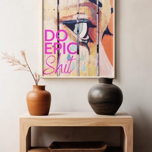 May include: A framed print with a colorful abstract painting of an eye with the text "DO EPIC SHIT" in pink letters. The print is hanging on a white wall above a wooden shelf with two vases and a woven basket.