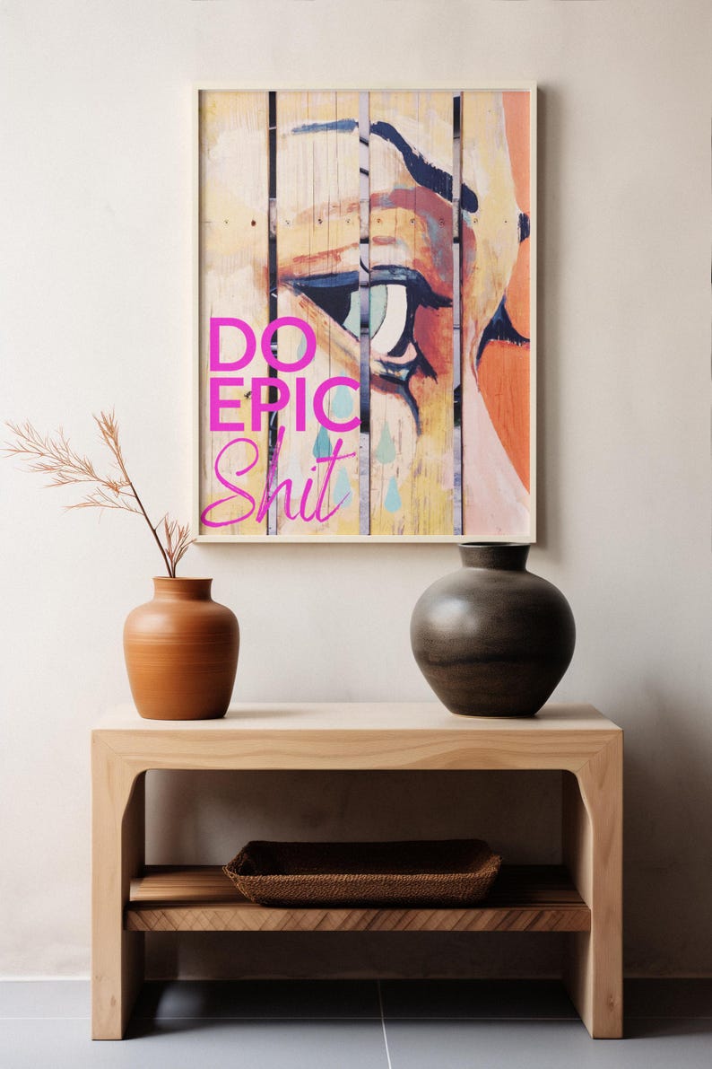 May include: A framed print with a colorful abstract painting of an eye with the text "DO EPIC SHIT" in pink letters. The print is hanging on a white wall above a wooden shelf with two vases and a woven basket.