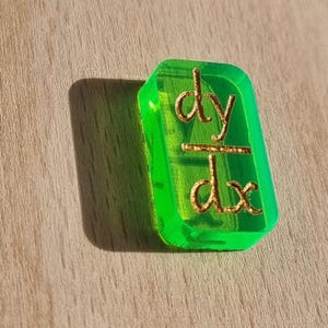 May include: A green plastic tile with the mathematical equation "dy/dx" written in gold lettering.