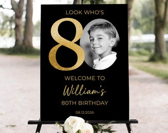 May include: A black sign with gold text and a photo of a young boy. The sign reads "Look Who's 80, Welcome to William's 80th Birthday, 08.13.2026". The sign is displayed on a black easel with white flowers.