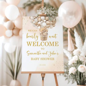 May include: A welcome sign for a baby shower, featuring the text "We can bearly wait WELCOME to Samantha and John's BABY SHOWER." The sign is decorated with a teddy bear in a basket and gold and white balloons. The sign is displayed on a wooden easel.