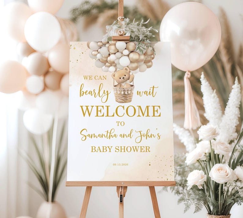 May include: A welcome sign for a baby shower, featuring the text "We can bearly wait WELCOME to Samantha and John's BABY SHOWER." The sign is decorated with a teddy bear in a basket and gold and white balloons. The sign is displayed on a wooden easel.