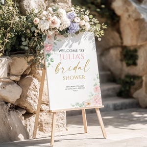 May include: A white sign on a wooden easel with the text "WELCOME TO JULIA'S bridal SHOWER" in gold script. Floral accents in pink, white, and green surround the text. The sign also includes the date "SEPTEMBER 13TH, 2024" and the phrase "60 DAYS UNTIL SHE SAYS 'I DO'."