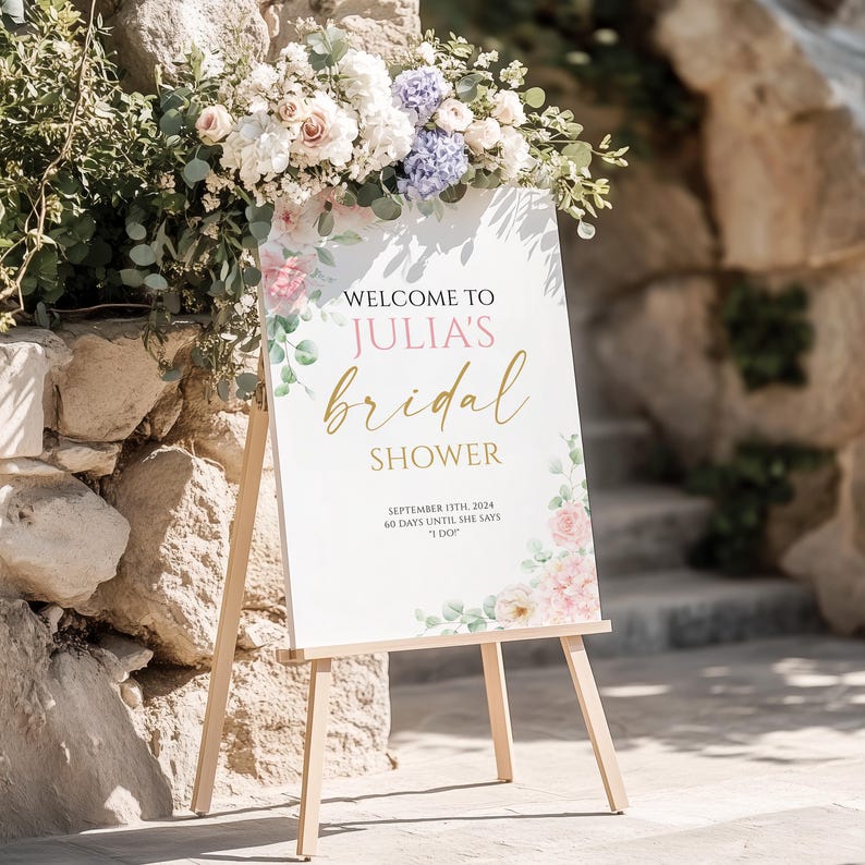 May include: A white sign on a wooden easel with the text "WELCOME TO JULIA'S bridal SHOWER" in gold script. Floral accents in pink, white, and green surround the text. The sign also includes the date "SEPTEMBER 13TH, 2024" and the phrase "60 DAYS UNTIL SHE SAYS 'I DO'."