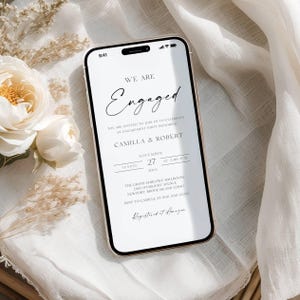Könnte beinhalten: Ein Smartphone zeigt eine Einladung zur Verlobungsfeier. Die Einladung lautet "We are Engaged" mit Details für Camilla & Robert, einschließlich Datum, Uhrzeit und Veranstaltungsort. Das Telefon liegt auf einer weißen Oberfläche mit floralen und Stoffakzenten.