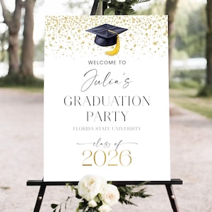 May include: A white graduation party sign with gold accents. The sign reads "Welcome to Julia's Graduation Party" with the Florida State University logo and "Class of 2026". A black graduation cap with a gold tassel is at the top.