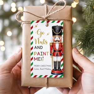 May include: A rectangular gift tag with a red and green striped border, featuring a nutcracker illustration and the text Go Nuts AND PAINT ME! MERRY CHRISTMAS! Love, Aunt Kaley. The tag is attached to a brown paper wrapped gift with twine.